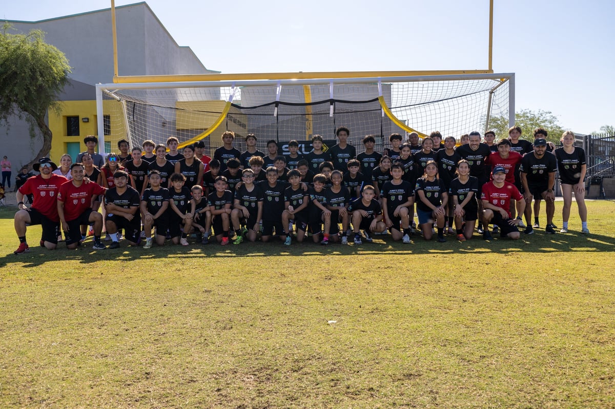 Large group of soccer players in red and black uniforms posing together on a grass field with a goal in the background